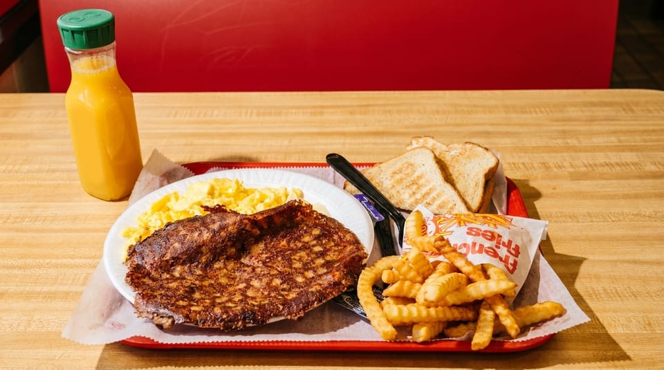 A breakfast tray with scrambled eggs, hash browns, toast, fries, and orange juice on a wooden table.