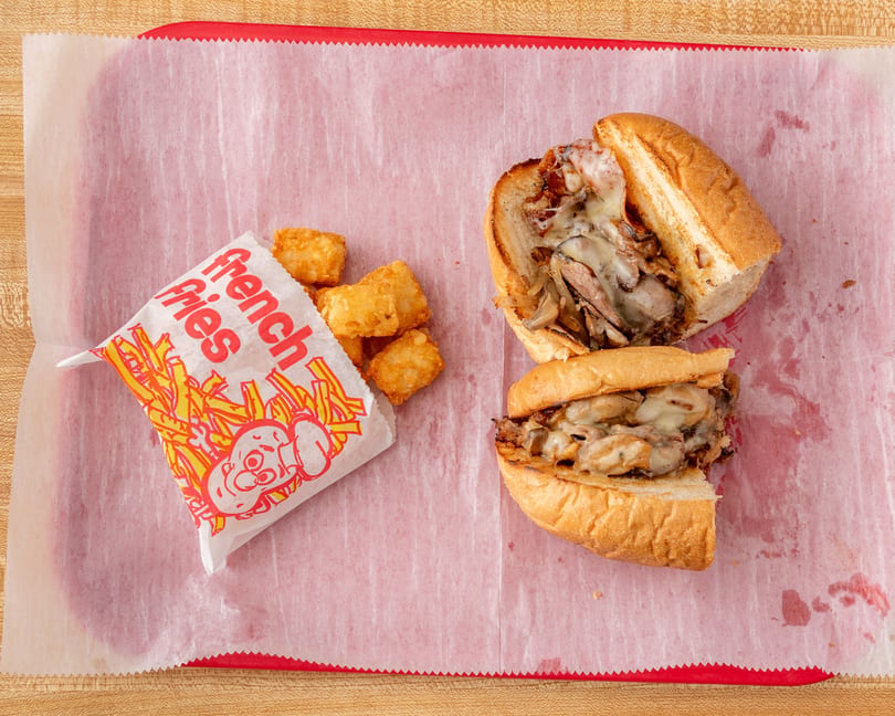 Two halves of a cheesesteak sandwich with mushrooms and onions, next to a bag of tater tots.
