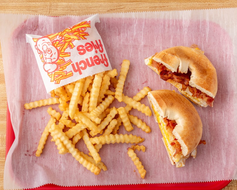 Crinkle-cut french fries spilling from a bag next to two bacon, egg, and cheese bagels.