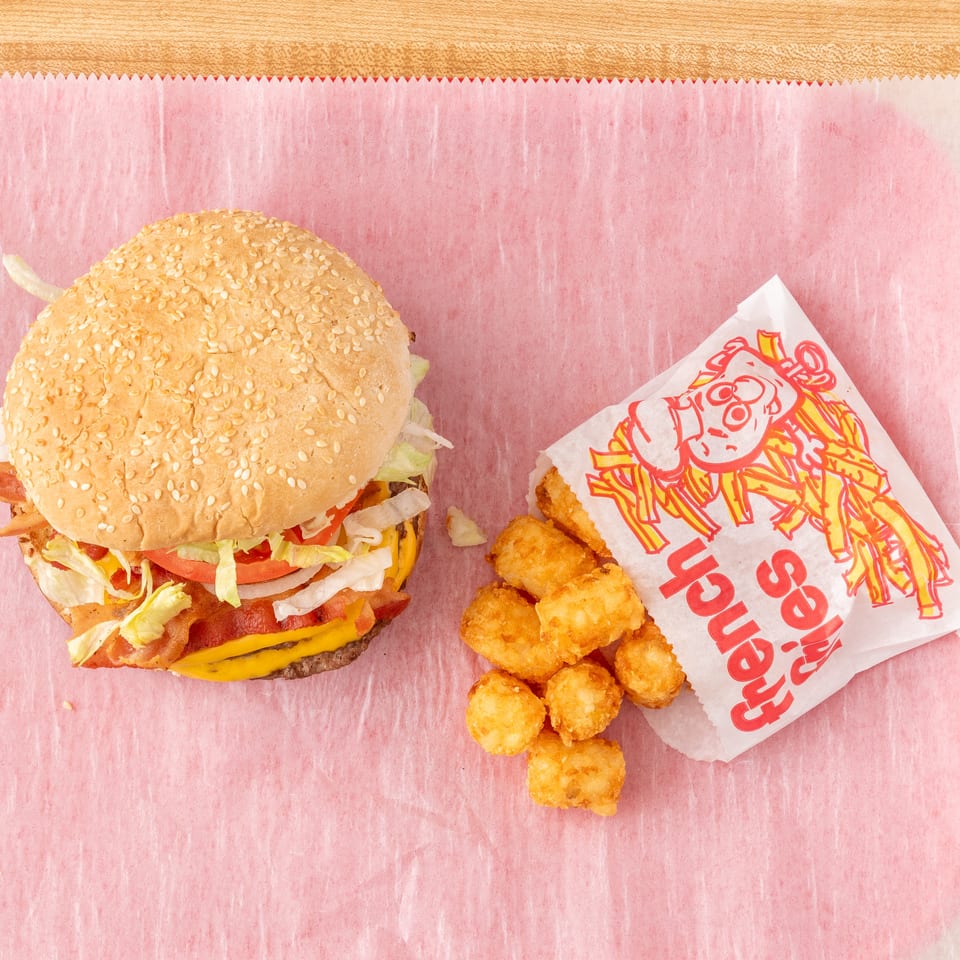 A bacon cheeseburger with lettuce, tomato, and onion, next to a bag of tater tots.