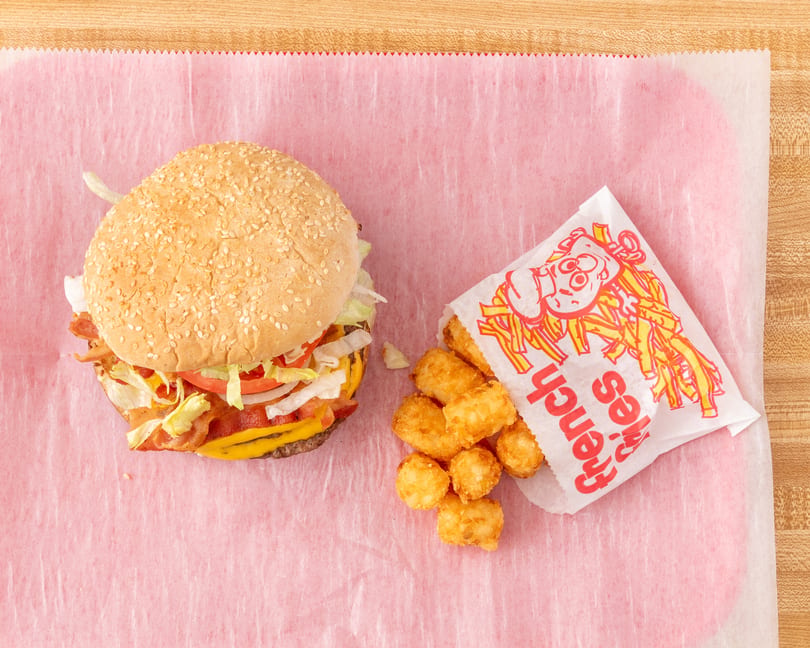 A bacon cheeseburger with lettuce, tomato, and onion, next to a bag of tater tots.