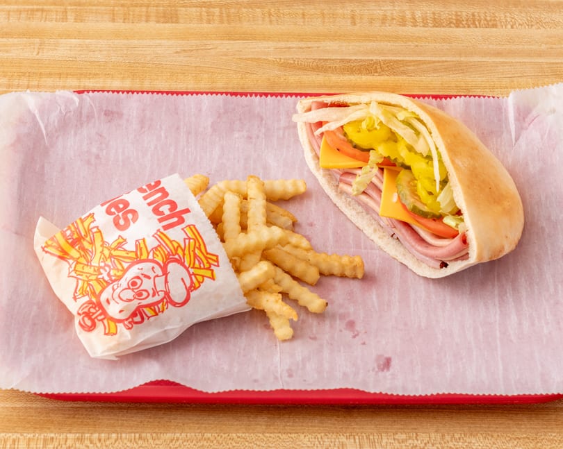 A pita sandwich with ham, cheese, and vegetables, next to crinkle-cut fries in a paper bag.