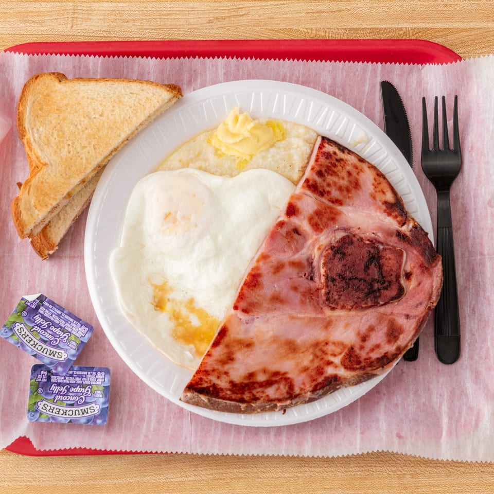 A breakfast platter with ham, fried egg, grits with butter, toast, and grape jelly packets.