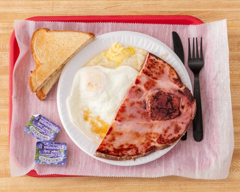 A breakfast platter with ham, fried egg, grits with butter, toast, and grape jelly packets.